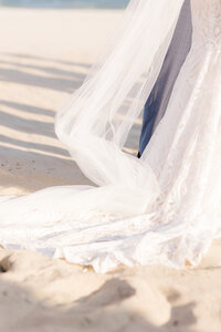 Close-up wedding detail of a bride’s lace gown and flowing veil trailing softly across the sand at a coastal wedding.