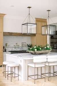 Bright organic kitchen with marble island, white stools, wood cabinetry, glass pendant lights, and a lush floral centerpiece creating an elegant, welcoming mood.