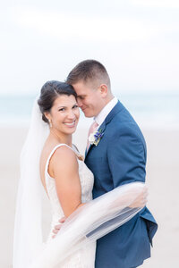 Bride and groom embracing on the beach with her veil wrapped softly around them during a coastal Delaware wedding.