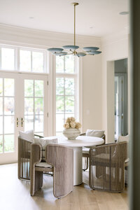 Bright, elegant dining area with a round white table, woven wicker chairs, floral centerpiece, and large glass doors overlooking greenery.
