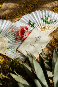 Wedding vow book, gold wedding rings and hand-painted floral fans styled with flowers and desert greenery.