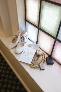 White Mary Jane heels, calligraphy wedding invitation, velvet ring box, and perfume arranged on a chapel windowsill bathed in stained-glass light