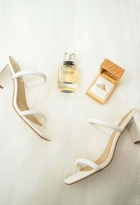 White shoes with a brown ring box that has a wedding ring inside. There is also a perfume bottle next to the other items. The background is plain white.