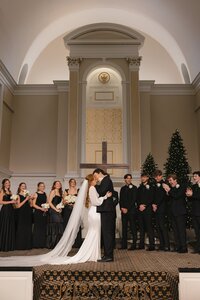 Bride and groom share their first kiss at the altar of Liberty University Chapel surrounded by a cheering wedding party at Christmas