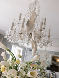 White bridal heels hanging from a crystal chandelier surrounded by wedding florals during bridal detail photography.