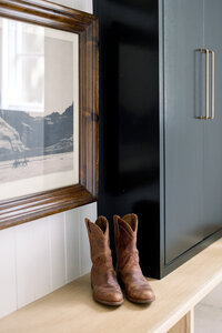 A pair of brown cowboy boots rests on a wood shelf beside framed mountain art and a dark cabinet, creating a warm, rustic interior.