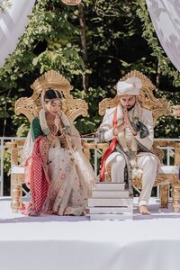 Indian bride and groom seated on ornate golden thrones with hands pressed in prayer during an outdoor mandap ceremony