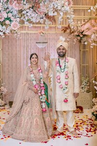 Radiant South Asian bride and groom in pink lehenga and ivory sherwani pose joyfully beneath a lush floral archway scattered with rose petals