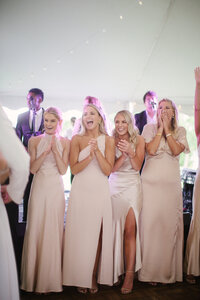 Bridesmaids in champagne colored dresses laugh and clap on the dance floor under a reception tent