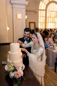 A laughing South Asian bride in gold-embroidered attire and groom in tuxedo joyfully cut their tiered wedding cake at a warmly lit DC reception