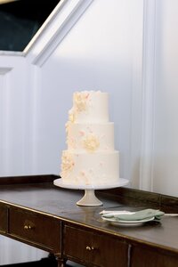 Elegant three-tier white wedding cake adorned with blush and cream sugar flowers displayed on a pedestal stand at a warm intimate reception
