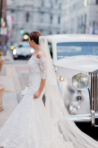 Bride in a long sleeve lace gown stands beside a vintage Rolls-Royce on a city street
