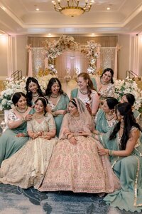 Radiant Indian bride in pink lehenga laughs with bridesmaids in sage sarees before an ornate floral mandap