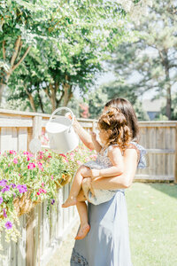 A mother holding her daughter while watering flowers in their backyard during an in home photography session in Wilmington, North Carolina
