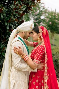 Indian bride in red lehenga and groom in ivory sherwani touch foreheads tenderly in a lush garden setting