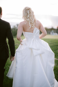 Bride's oversized bow dress detail seen from behind as she walks beside the groom
