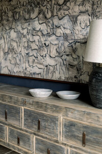 Weathered wood sideboard with two white bowls, a dark lamp, and a large expressive charcoal sketch of figures, creating a rustic, moody mood.