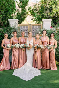 Bride in a lace wedding gown surrounded by bridesmaids in blush satin dresses holding floral bouquets.