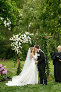 Bride and groom kissing at alter of their wedding with priest in the background with a lot of greenery around them at the Stevens Osgood Estate in Massachusettes.