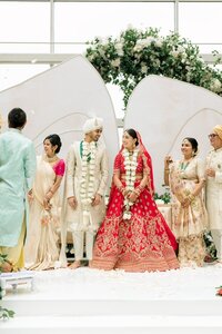 Bride in red embroidered lehenga and groom in ivory sherwani exchange garlands joyfully beneath a lush white floral archway