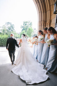 Bride and groom walk hand in hand past bridesmaids in pale blue gowns under a stone archway