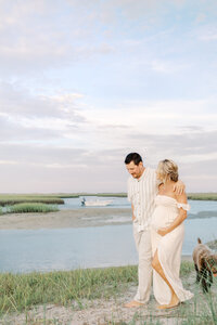 A couple walking along the beach with their dog in the backdrop during their maternity photography session at Wrightsville Beach, North Carolina