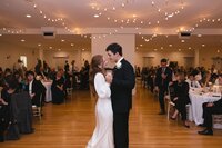 Bride and groom share a tender first dance on a warm ballroom floor beneath twinkling string lights as guests look on