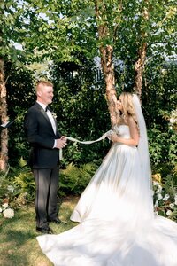 Smiling bride and groom hold a braided rope during an outdoor unity ceremony beneath sunlit birch trees