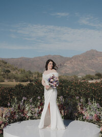 Bride holding a colorful bouquet in front of desert landscaping and Arizona mountain views.