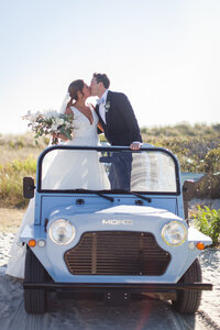 Bride and groom kiss while standing in a vintage blue beach vehicle near the dunes