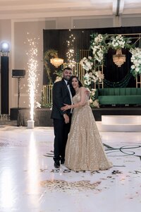 Bride in a gold sequined lehenga and groom in a tuxedo embrace on a monogrammed dance floor amid sparkling pyrotechnics