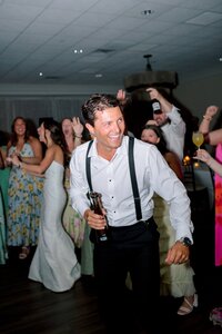 Grinning groom in white shirt and suspenders dances energetically on the reception floor surrounded by cheering guests