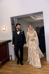 Smiling South Asian bride in gold embroidered lehenga and groom in black tuxedo walk hand-in-hand through a warm indoor archway