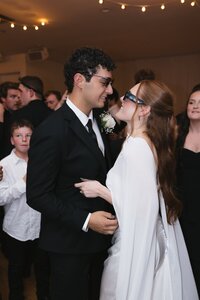 Smiling bride in white cape gown and groom in black suit wearing sunglasses share a joyful dance at their intimate wedding reception