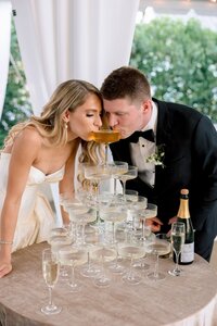 Bride and groom lean in cheek-to-cheek sipping from the top coupe of an elegant champagne tower at their tented Northern Virginia reception