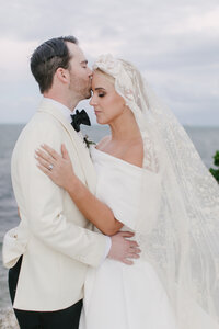 Groom kisses bride's forehead by the ocean as her lace veil blows in the wind