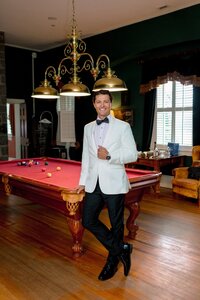 Confident groom in a white tuxedo jacket leans against a red billiard table in an elegant dark-walled game room