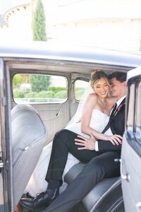 A bride and groom sit closely together in the back seat of a vintage car, smiling and embracing, dressed in wedding attire. Sunlight fills the car, highlighting their happiness.