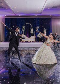 Indian bride in embroidered lehenga and groom in black suit dancing exuberantly on a sparkling ballroom floor beneath purple uplighting