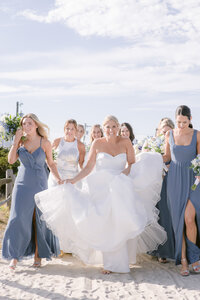 Bride laughs while walking on the beach surrounded by bridesmaids in dusty blue gowns