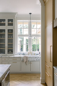 Bright, luxury kitchen with marble island, glass-front cabinets, brass fixtures, pendant light, and sunlit windows creating a calm tone.