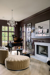 Moody dark-paneled living room with grand piano, chandelier, fireplace, framed art, and curved velvet seating; warm, sophisticated mood.