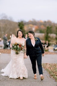 A bride wearing a white lace dress holding a bouquet of yellow, red and orange flowers walks hand in hand with her wife who is wearing a navy suit and small boutonniere of greenery