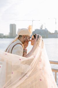 South Asian bride and groom in ivory wedding attire share a tender nose-touch moment on a sunlit Norfolk waterfront, her embroidered dupatta billowing in the breeze