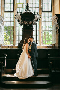 Bride and groom embracing beside a large arched window inside their wedding venue