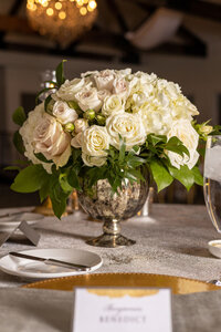 Elegant wedding centerpiece of white hydrangeas, roses and greenery arranged in a silver vase.