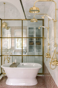 Luxury bathroom with a white freestanding bathtub, brass fixtures and rainfall shower, glass doors, marble walls, and warm natural light.
