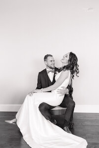 A bride in a long gown sits on a grooms lap. Both are smiling and dressed formally. They pose against a plain wall in a light, airy room with wooden floors.