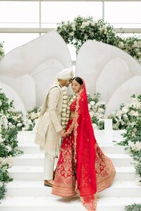 Indian bride in crimson gold lehenga and groom in ivory sherwani share an intimate forehead touch at a white floral mandap