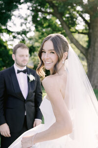 Bride glances back at the camera smiling as her groom stands beneath a shaded tree canopy